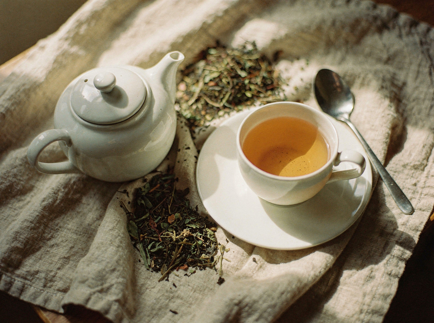 A white teacup on its saucer beside a small teapot and a scattering of dry loose leaf tea on linen
