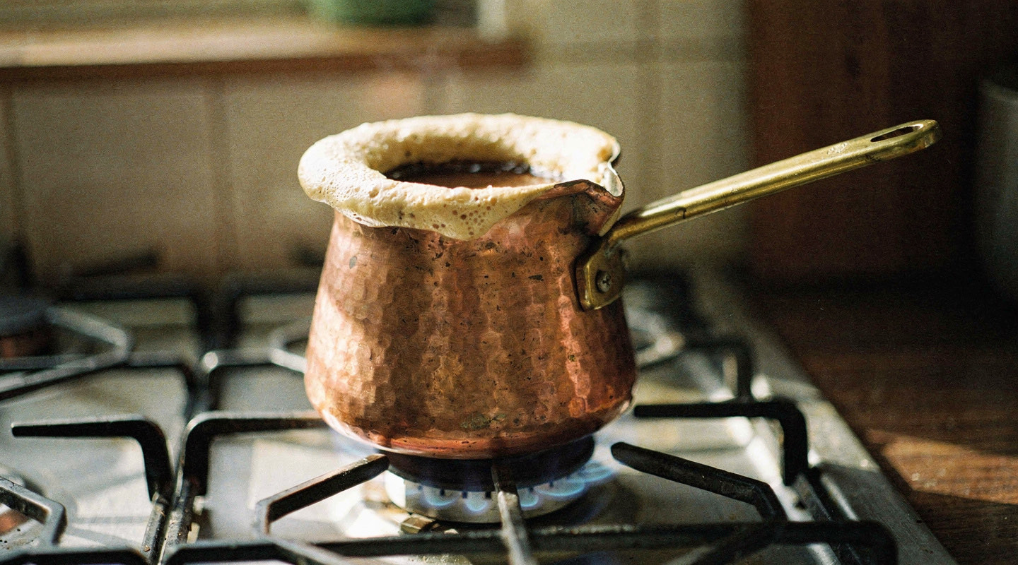 A small copper cezve on a low flame with a thick pale ring of foam rising toward the rim