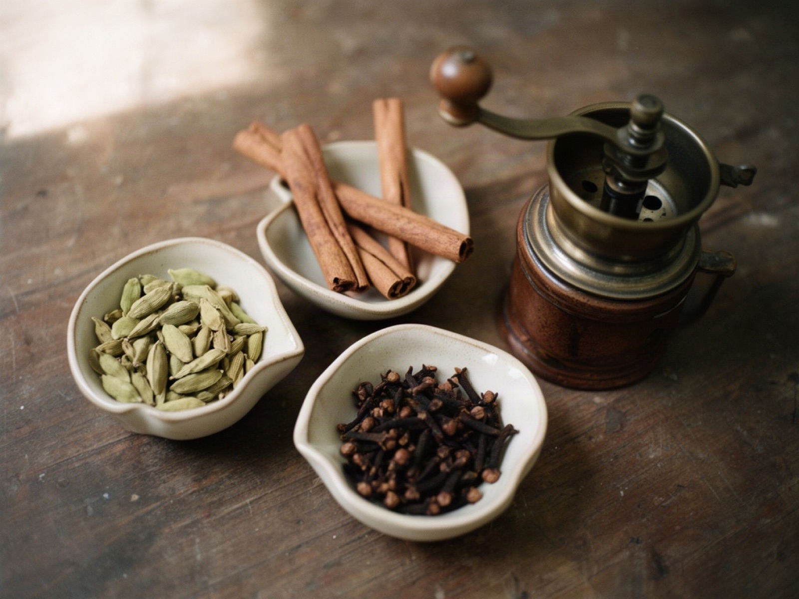Small bowls of cardamom pods, cinnamon sticks and cloves beside a brass coffee grinder