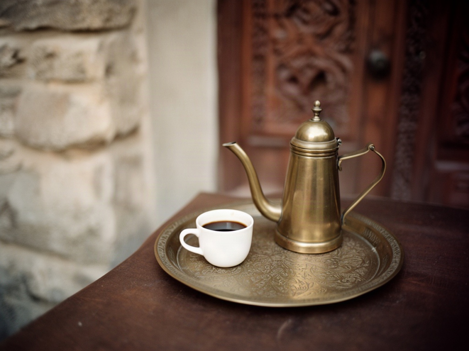 A brass coffee pot and small white cup on an engraved tray in front of a carved wooden door