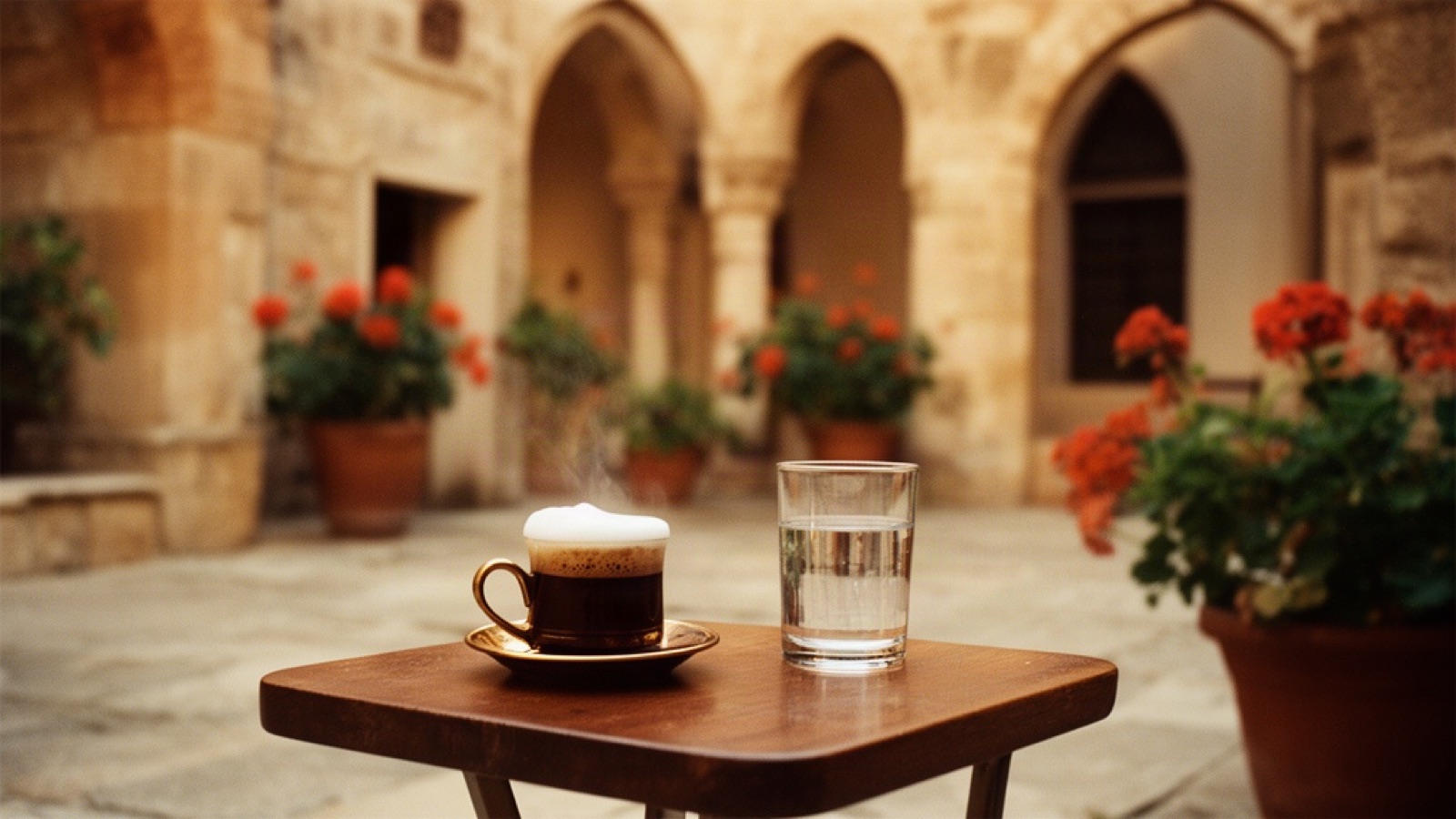 A foamy Turkish coffee and a glass of water on a small wooden table in a stone courtyard